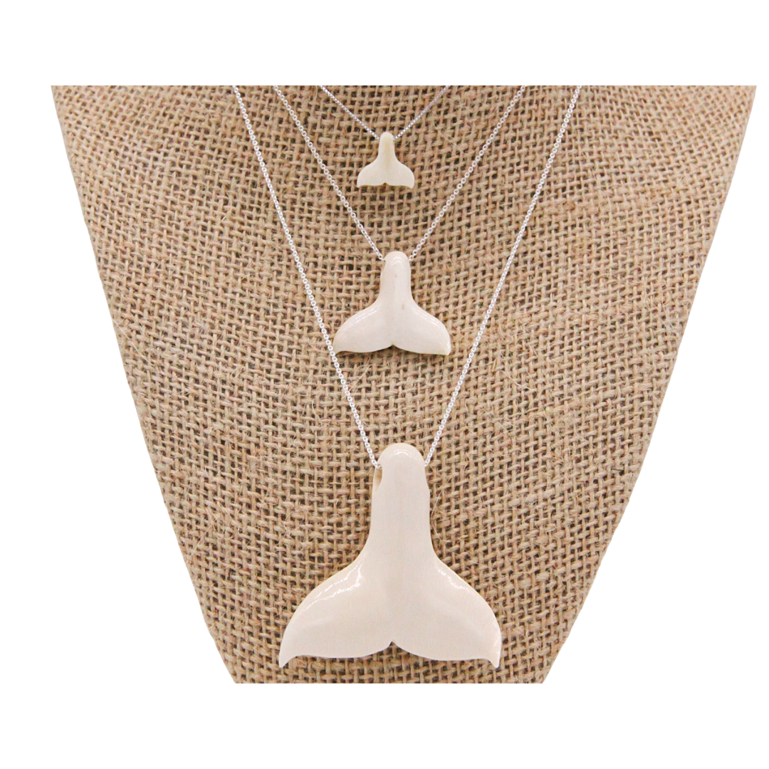 Three whale tail pendants from small to big, lay across a tan fabric.