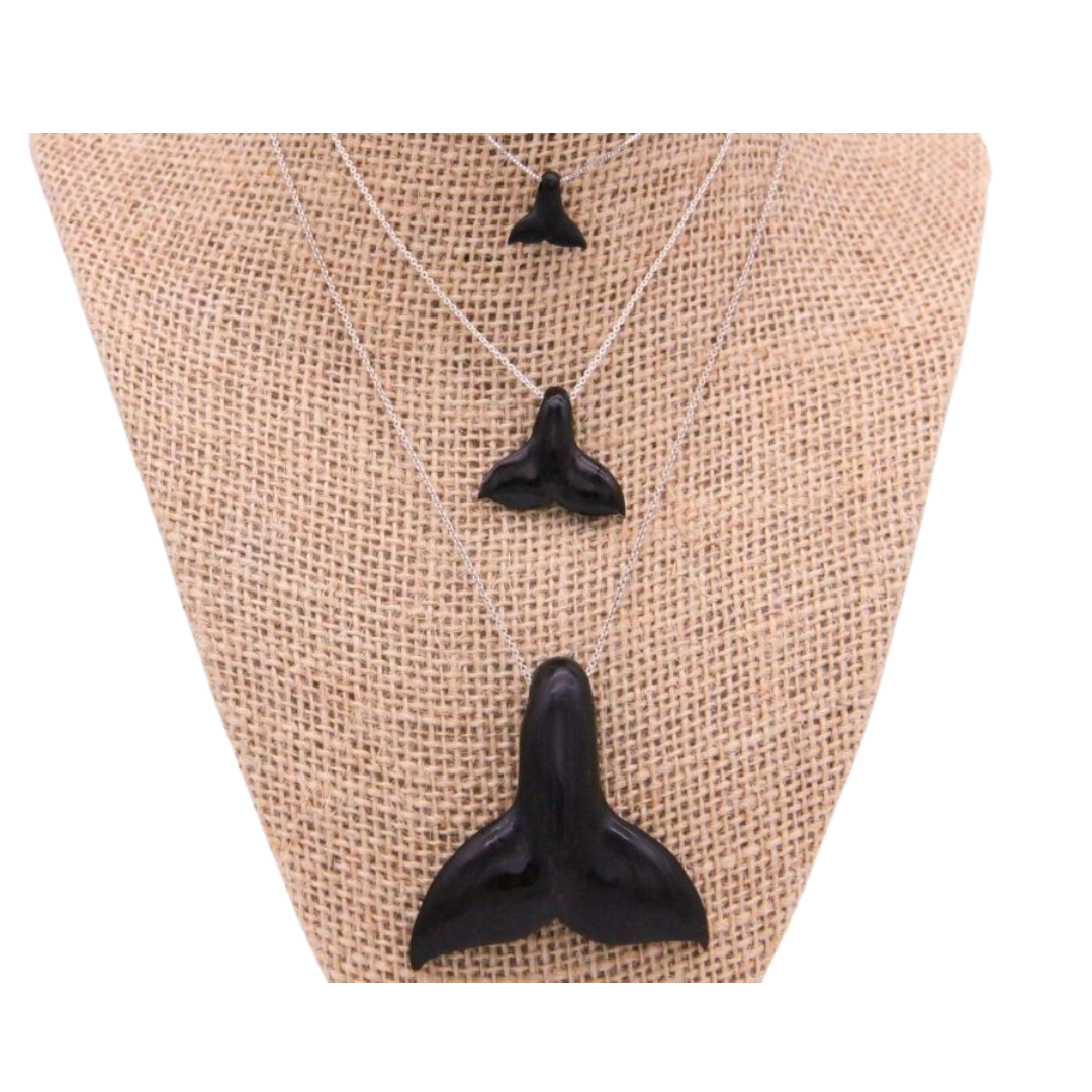 Three whale tail pendants on necklaces, in varying sizes, displayed on a burlap surface. The pendants are black in color, resembling the appearance of whale teeth.
