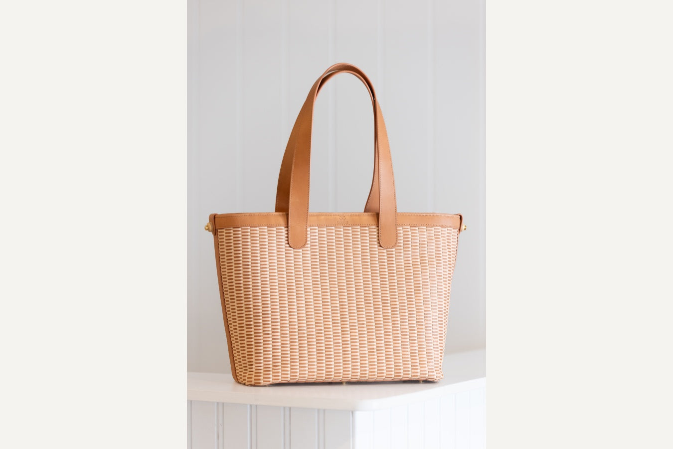 A woven tote bag with tan leather handles and trim, displayed on a white pedestal against a white paneled background.