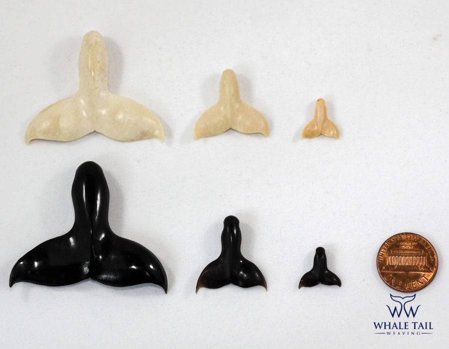 The fossil and ebony whale tail pendants sit side by side, varying in size as they are compared to a penny.